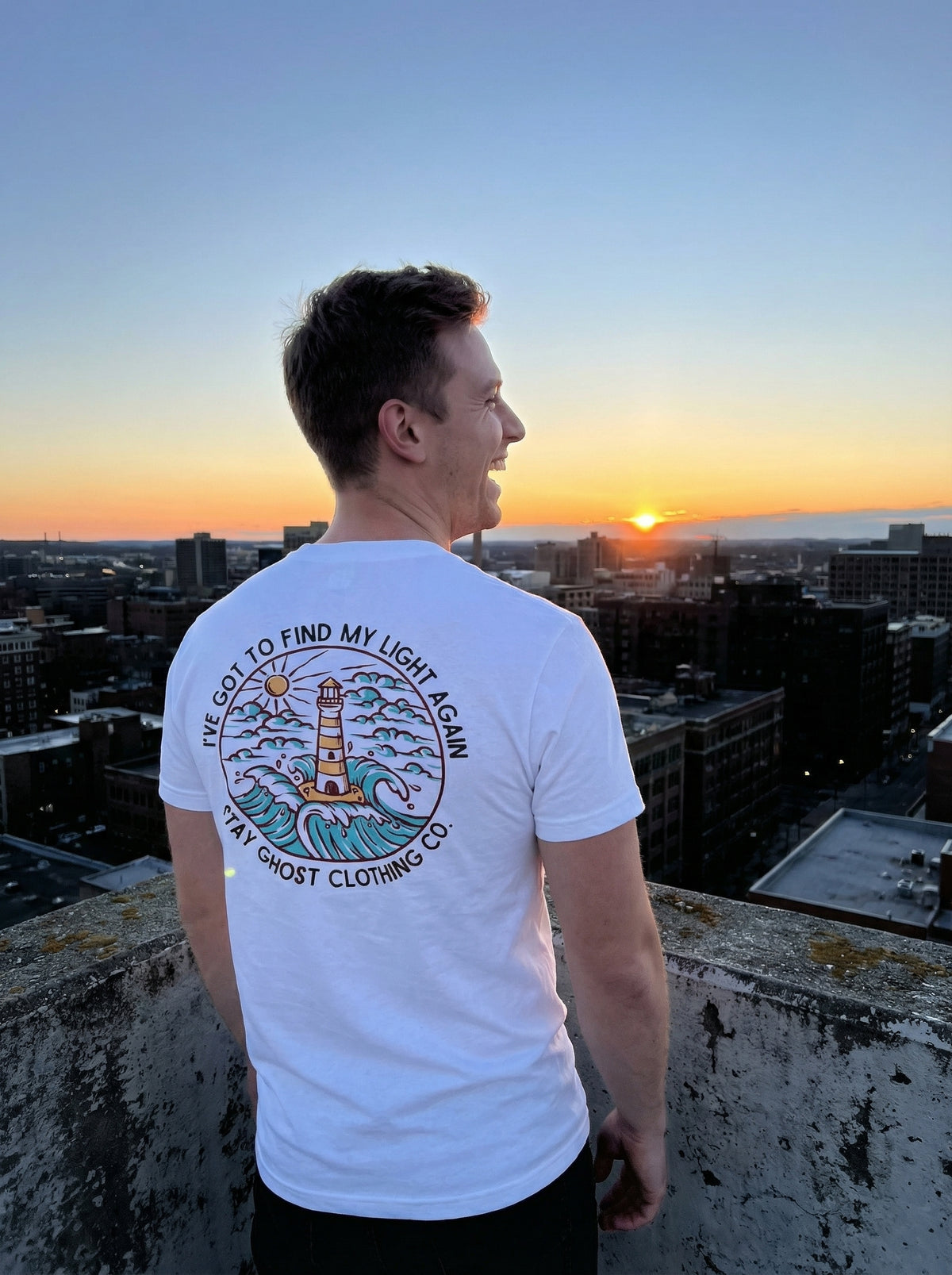 Person wearing a white t-shirt with a design and text, standing on a rooftop overlooking a cityscape at sunset.